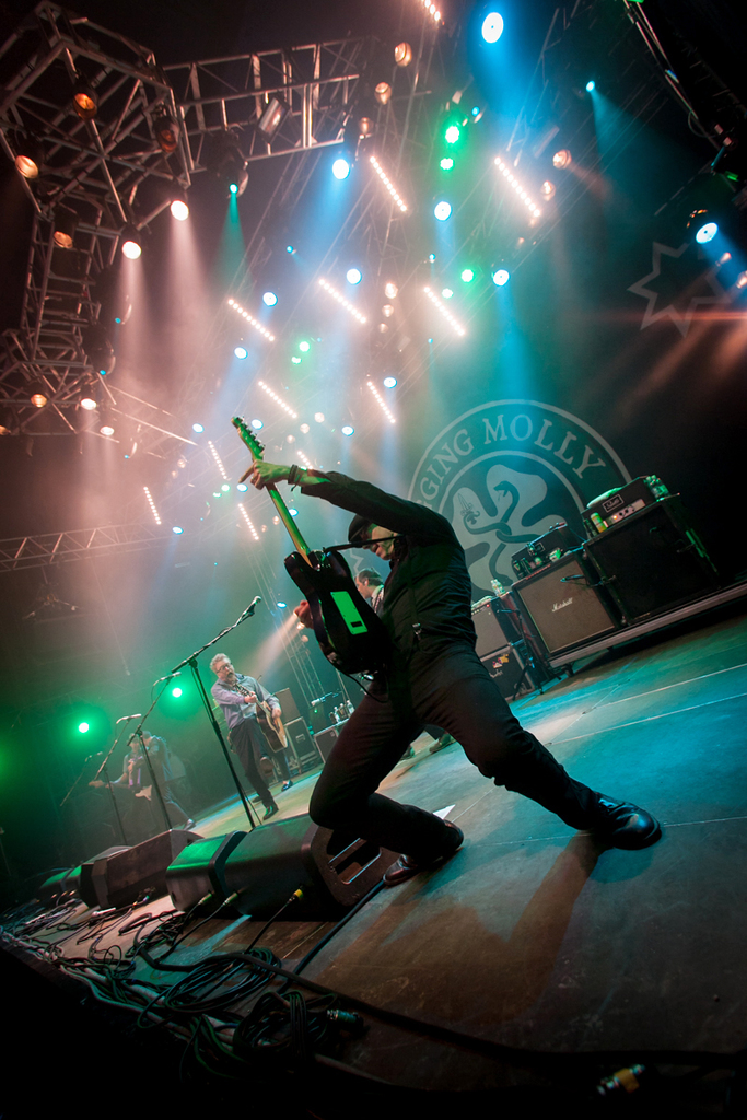 Ein musikalisches Konzert mit drei Künstlern auf der Bühne: zwei Gitarristen rechts mit Mikrofonen davor, ein Hintergrund mit dem Wort "Molly" und zwei Schlangenlogos sowie Scheinwerfer oben.