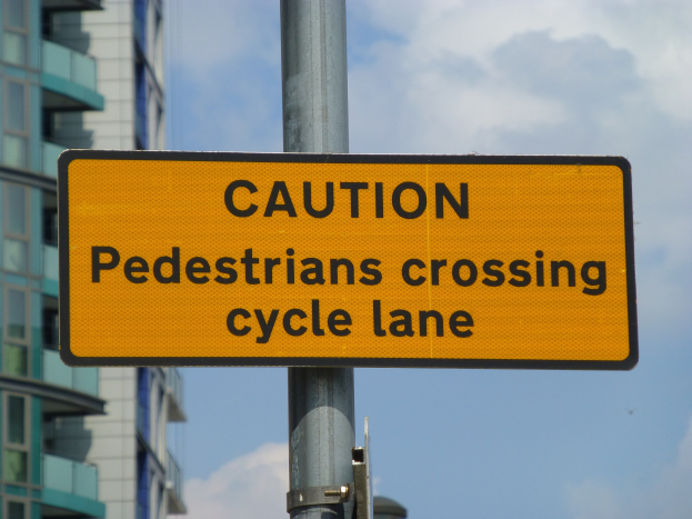 Gelbes Warnschild an einem Pfosten mit der Aufschrift "Fußgänger überqueren Fahrradspur" vor einem Gebäude mit einem klaren blauen Himmel.
