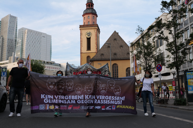 Eine Gruppe von Menschen mit Masken, die eine Straße entlanggehen und eine Fahne mit Text und Bildern halten, mit einem geparkten Auto auf der linken Seite, Gebäuden, Bäumen, Schildern, Pfählen, einem Kirchturm und einem klaren blauen Himmel im Hintergrund.