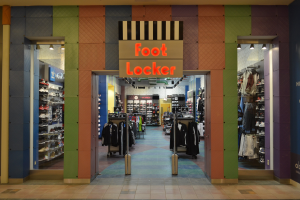 Innenansicht eines Foot Locker-Schuhgeschäfts mit Kleidung an Bügeln und Ständern unter Deckenbeleuchtung, wobei der Ladenboden sichtbar ist.