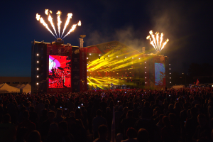 Eine große Menschenmenge auf einem Musikfestival mit einer hell erleuchteten Bühne, die Künstler und Banner zeigt, Zelte links und Feuerwerk am dunklen Himmel.