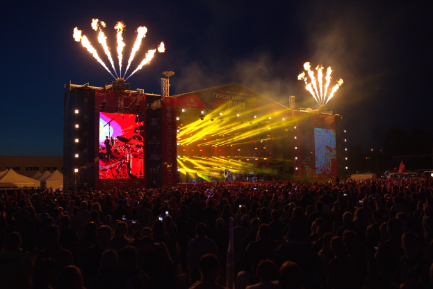 Eine große Menschenmenge auf einem Musikfestival mit einer hell erleuchteten Bühne, die Künstler und Banner zeigt, Zelte links und Feuerwerk am dunklen Himmel.