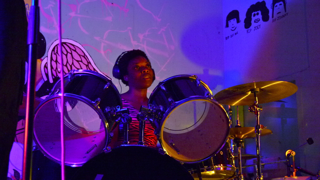 Eine Frau, die Musikinstrumente auf einer B├╝hne spielt, mit einem Mikrofonstativ und einer Person im Hintergrund.