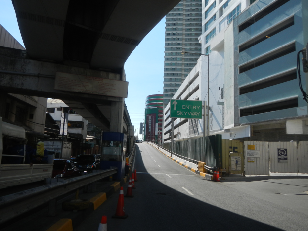 Eine Stadtstraße mit Absperrgittern am Straßenrand, Fahrzeuge auf der Straße, eine Brücke links, Gebäude mit Namensschildern auf beiden Seiten, Laternenpfähle und ein Himmel im Hintergrund.