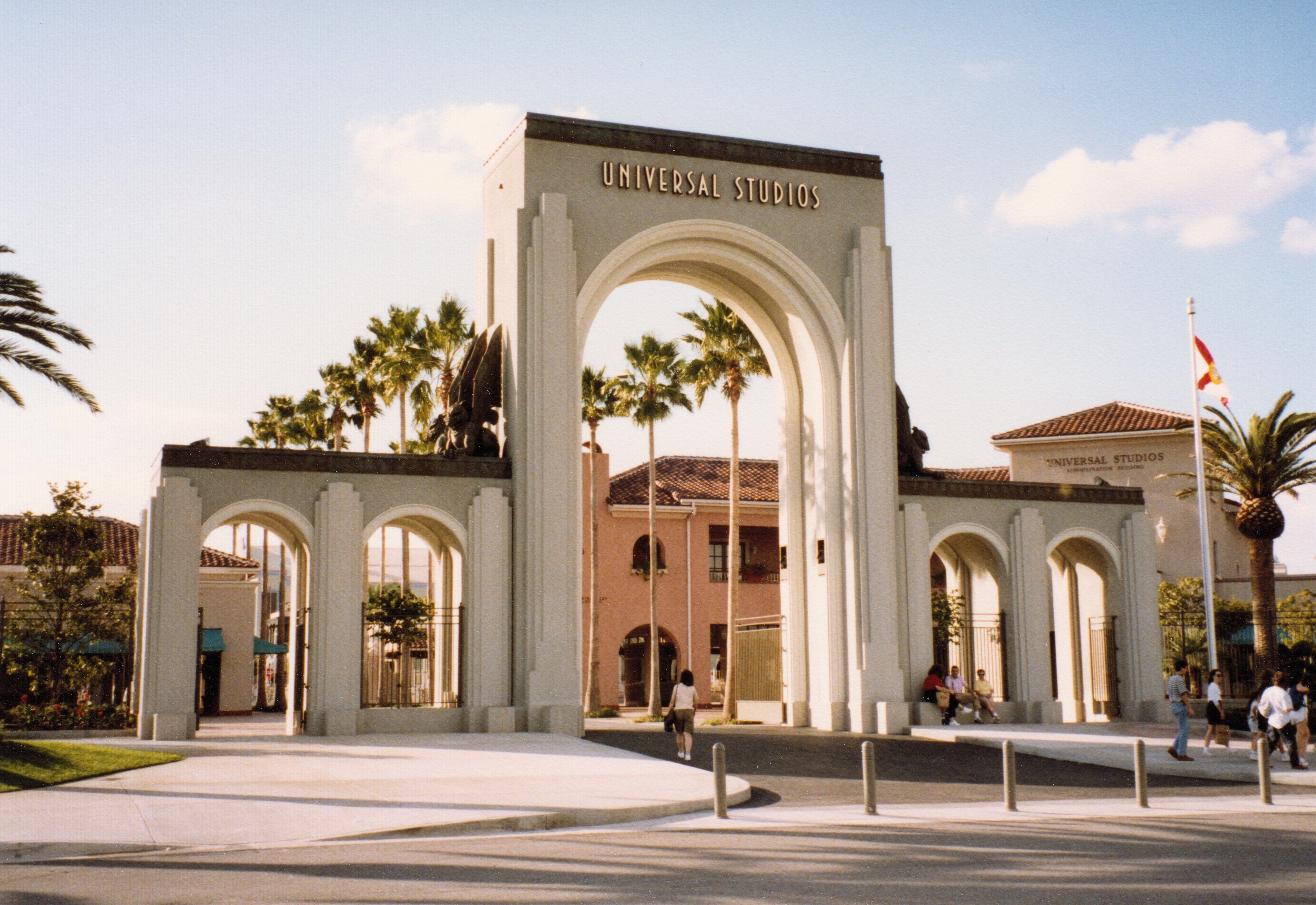 Eingang zu Universal Studios Hollywood mit einem Bogen mit Text, einer Statue, Menschen, umgeben von Grün, Gebäuden, einem Fahnenmast mit einer Flagge und einem bewölkten Himmel.