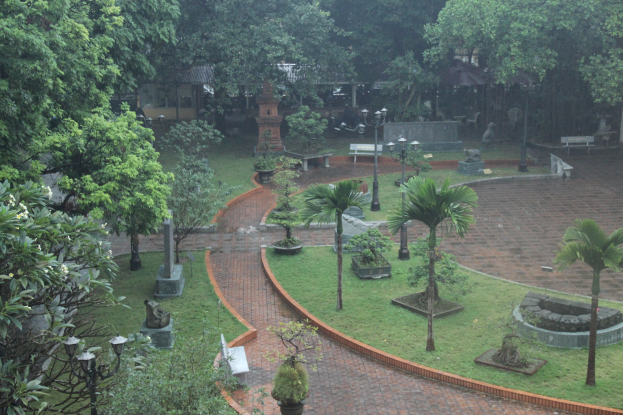 Ein grüner Park mit verschiedenen Bäumen, Pflanzen, Gras, Laternen, Bänken und Skulpturen, mit einem Gebäude im Hintergrund.