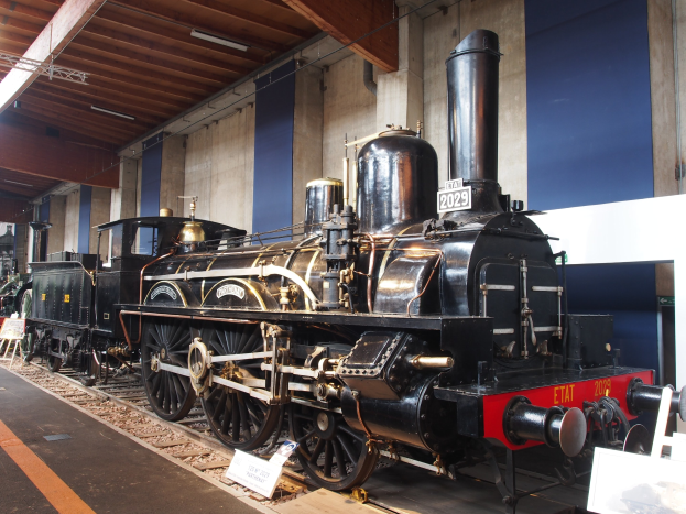 Alte Dampflokomotive in einem Museums-Setting mit umgebenen Eisenbahnschienen, Informations-Tafeln und anderen Ausstellungsstücken.