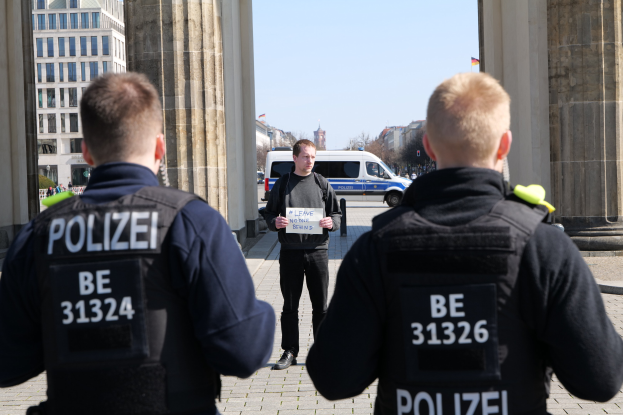 Drei Polizeibeamte stehen vor einem Gebäude, mit einem Mann in der Mitte, der ein Papier hält, flankiert von Säulen und umgeben von Fahrzeugen, Menschen, Bäumen und Gebäuden unter dem Himmel.