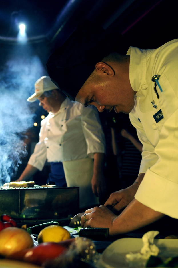 Zwei Köche in weißen Uniformen und Mützen kochen auf einem Grill in einer Küche, mit Gemüse auf dem Tisch und Lichtern im Hintergrund.