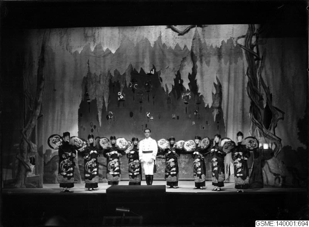 Schwarzes und weißes Foto von Darstellern auf der Bühne in traditionellen japanischen Kimonos, die Gegenstände halten, mit einer gemalten Kulisse und Text, der "Kimono-Show in Tokyo, Japan" lautet.