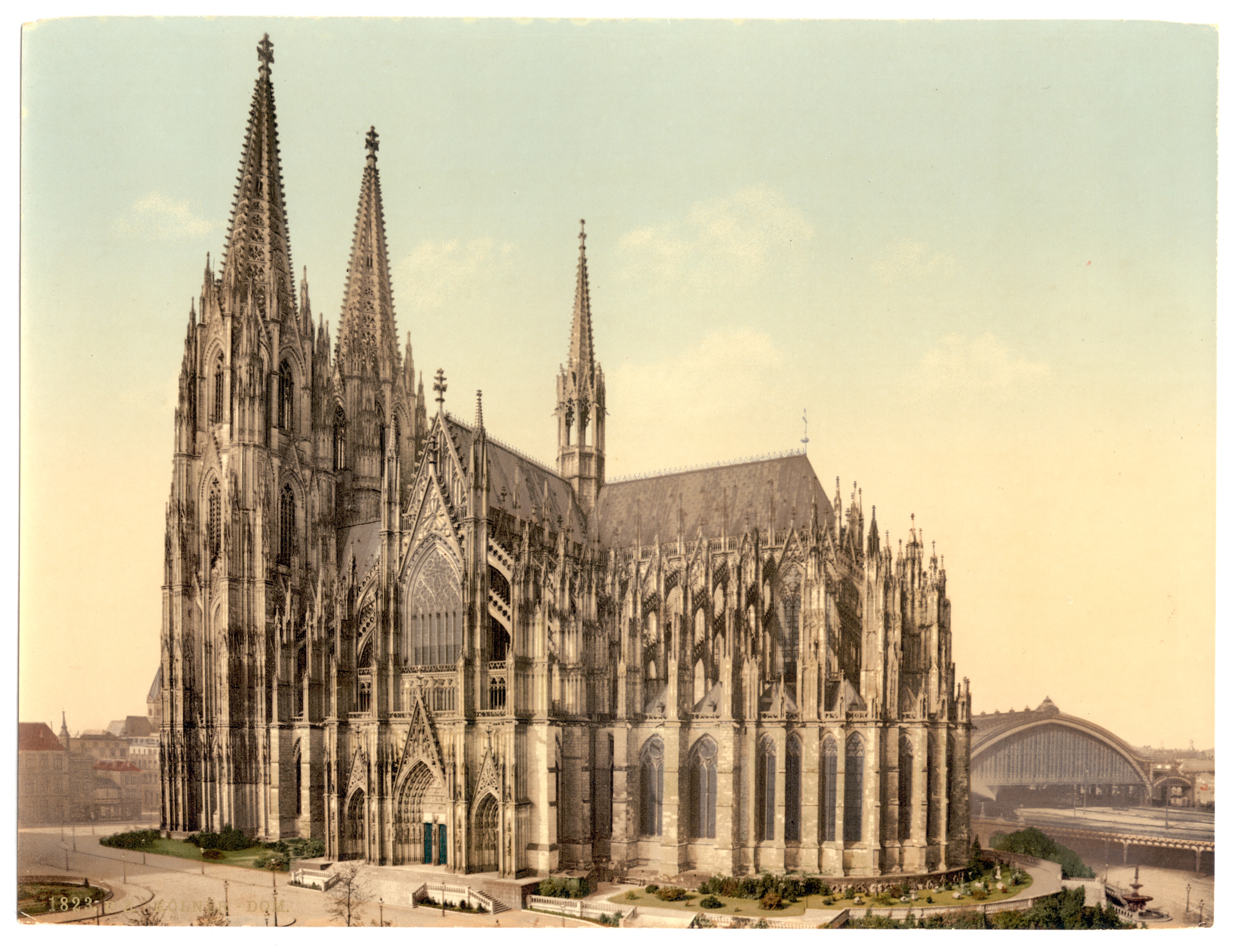 Ein altes Foto der Kölner Dom in Deutschland, mit seinen Spitzen, die sich bis in den Himmel erstrecken, umgeben von üppiger Vegetation, einer Brücke auf der rechten Seite, Fahrzeugen auf der Straße und anderen Gebäuden im Hintergrund unter einem klaren blauen Himmel.