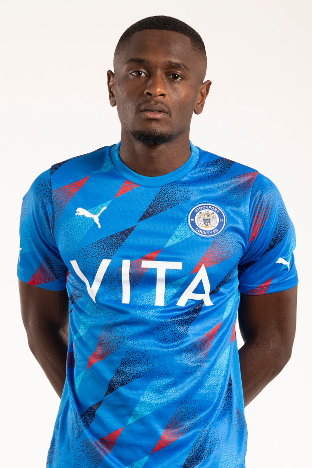 Ein Mann in einem blauen Leicester City Away-Shirt von der Saison 2018-19, der entspannt mit den Händen in den Taschen und einem leichten Lächeln dasteht.