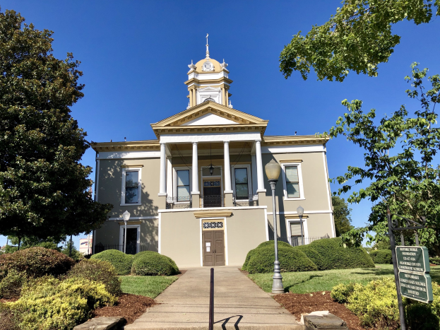 Großes Gebäude mit einem Uhrenturm, umgeben von Fenstern, Türen, Säulen, Geländern und einem Dach, das als das alte Rathaus bezeichnet wird, das kürzlich wiedereröffnet wurde, mit Straßenlaternen, Straßenschildern, Pflanzen, Gras, Baumrinde, Bäumen und einem bewölkten Himmel.