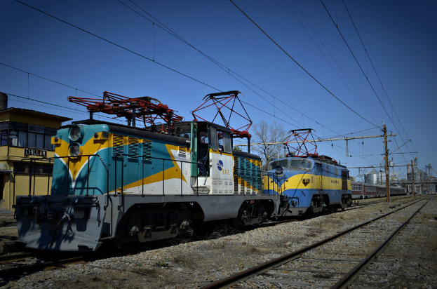 Zwei Zugmaschinen auf einem linken Gleis neben einem Gebäude mit einer schwarzen Namensschild, mit Strommasten, Drähten, einem Baum und Gebäuden im Hintergrund unter einem klaren Himmel.