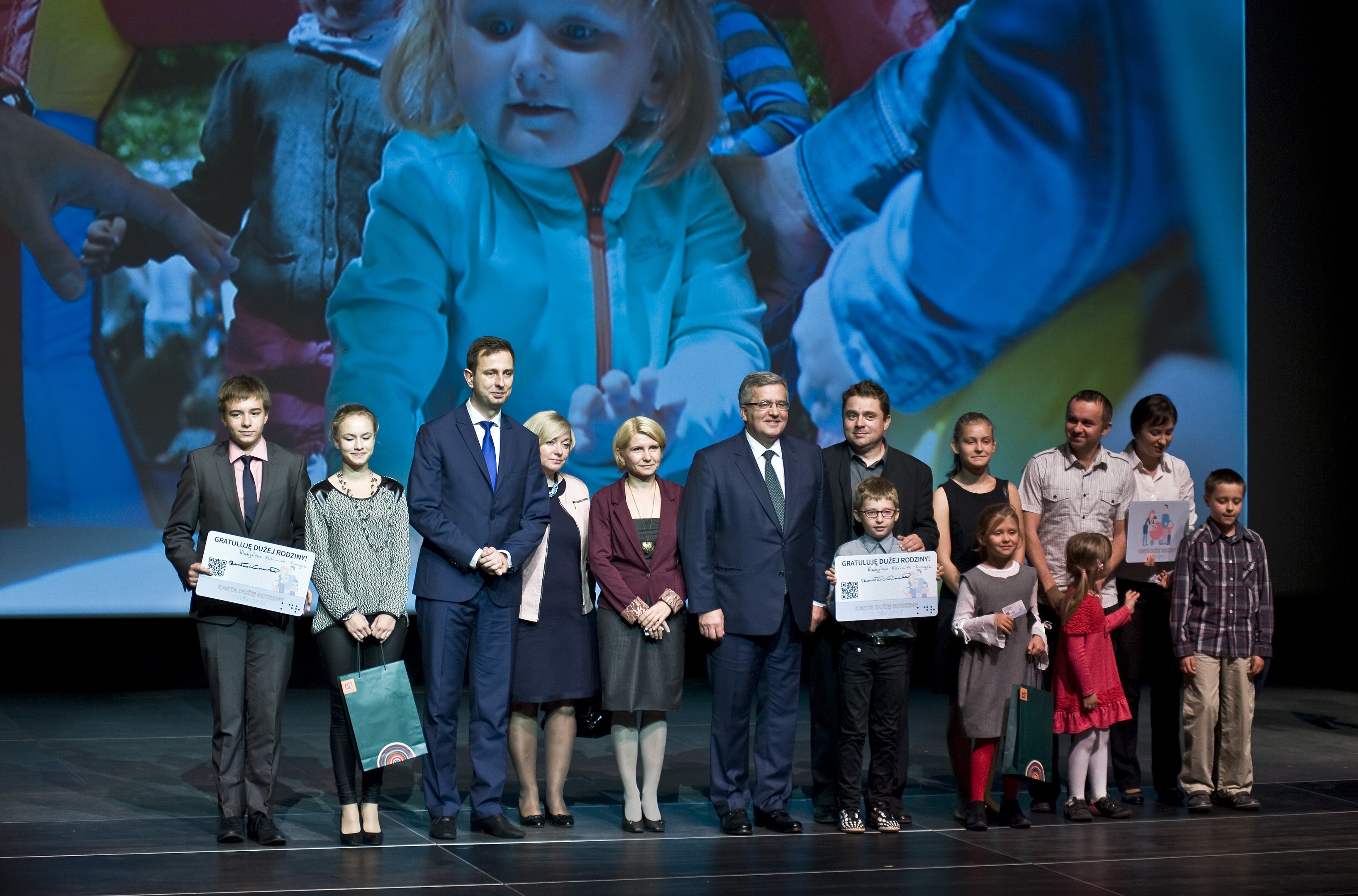 Eine Gruppe von Menschen auf einer Bühne mit Taschen und Urkunden, im Hintergrund ein Bildschirm mit einem Kind und anderen