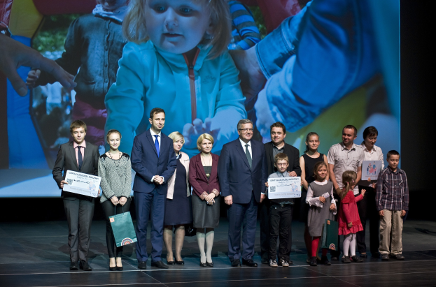 Eine Gruppe von Menschen auf einer Bühne mit Taschen und Urkunden, im Hintergrund ein Bildschirm mit einem Kind und anderen