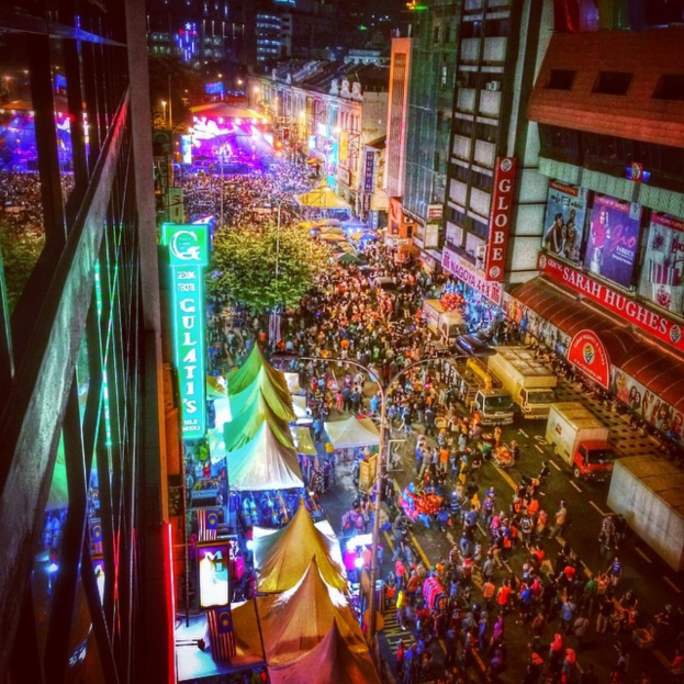 Eine lebhafte Karnevalszenen mit einer großen Menge auf der Straße, umgeben von Zelten, Geschäften, Gebäuden und Bannern, mit hell beleuchteten Bereichen im Hintergrund.