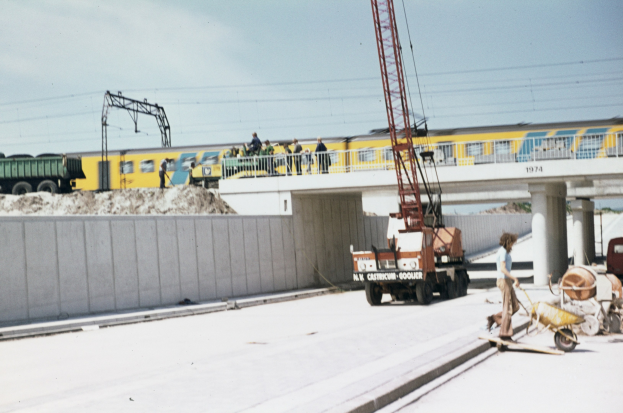 Baustelle mit einem Zug auf Gleisen, eine Brücke mit Säulen und Menschen, eine Person mit einem Handwagen, Fahrzeuge auf einer Straße, eine Wand, Pfosten, Drähte und eine bewölkte Landschaft.