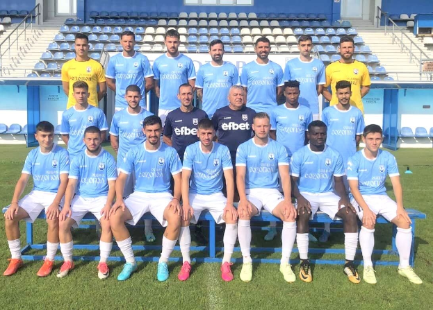 Eine Gruppe von Männern, einige sitzen auf Bänken und andere stehen auf einem Fußballfeld, mit Tribünen und Gebäuden im Hintergrund, wahrscheinlich feiern sie einen Mannschaftssieg.