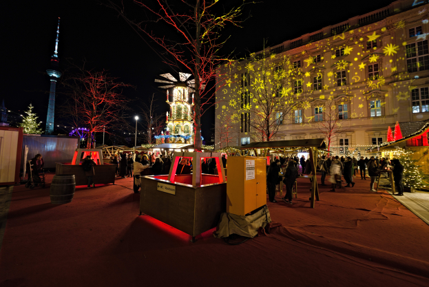Ein lebhaftes Weihnachtsmarkt in Berlin, Deutschland, mit Menschen um beleuchtete Stände, Bäume, Gebäude, Laternenmäste und einen Turm unter einem dunklen Himmel.