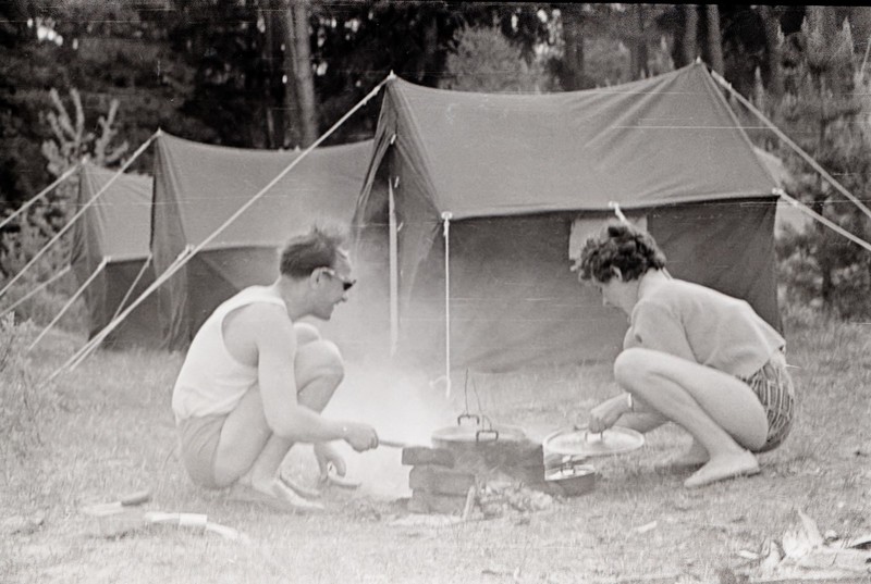 Ein Mann und eine Frau sitzen um ein Lagerfeuer vor einem Zelt, umgeben von Bäumen in einem Schwarz-Weiß-Bild.