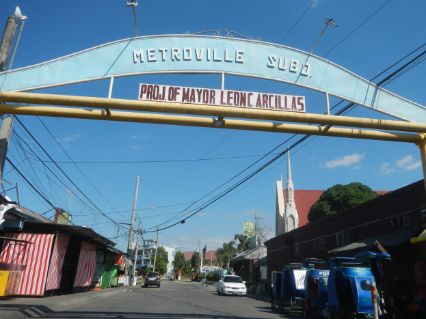 Bogen mit "Metroville Subo - Projekt des Bürgermeisters Leon Arcillas" Text, Fahrzeuge auf der Straße, Schuppen links, Strommasten mit Drähten, Bäume und Gebäude im Hintergrund, bewölkter Himmel oben.