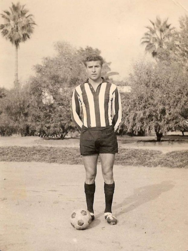 Schwarzes und weißes Foto eines älteren Mannes, der einen Fußball hält, draußen mit Bäumen und Himmel im Hintergrund.