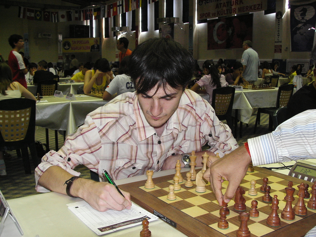 Eine Hand eines Mannes bewegt ein Schachspiel auf einem Brett, während er mit einem Stift schreibt, mit spielenden Menschen im Hintergrund.