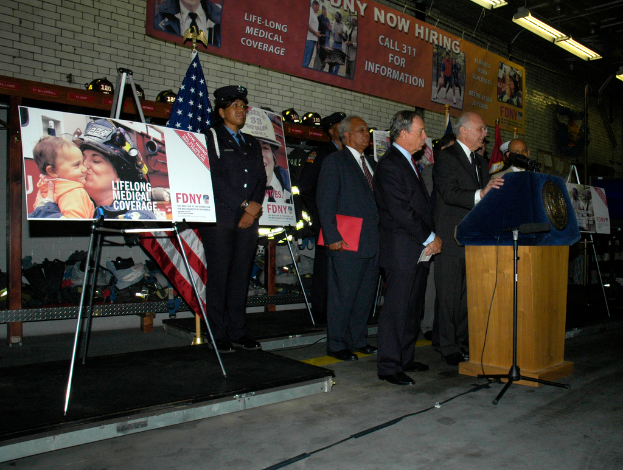 Eine Gruppe von Menschen steht in der Nähe eines Podiums mit Mikrofonen, umgeben von Tafeln auf Ständern, Fahnen und Tafeln an der Wand, alles unter hellen Scheinwerfern.