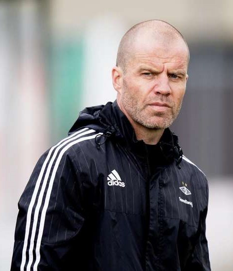 Ein kahlköpfiger Mann in einer blauen Jacke mit weißen Streifen und einem weißen Kragen steht auf einem Fußballfeld, die Hände verschränkt, mit einem ernsten Gesichtsausdruck und einem unscharfen Hintergrund.