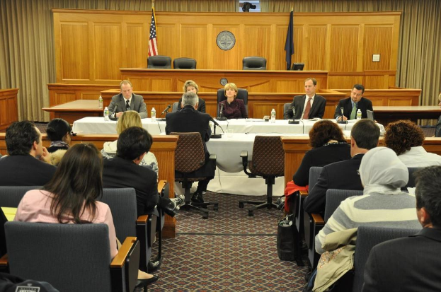Ein Versammlungsraum mit auf Stühlen sitzenden Menschen, zwei ausländische Flaggen, ein oranger Schreibtisch mit Wasserflaschen und einem Mikrofon.