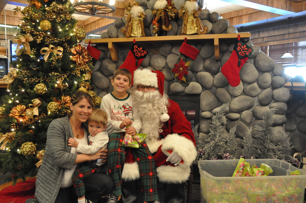 Eine Frau, Kinder und ein Mann in Santa-Klaus-Kostüm mit Brille und Handschuhen stehen neben einem geschmückten Weihnachtsbaum, einer Wand mit Steinen, Spielzeugen, Dekorationen und einem Korb mit Gegenständen, mit Lichtern im Hintergrund.