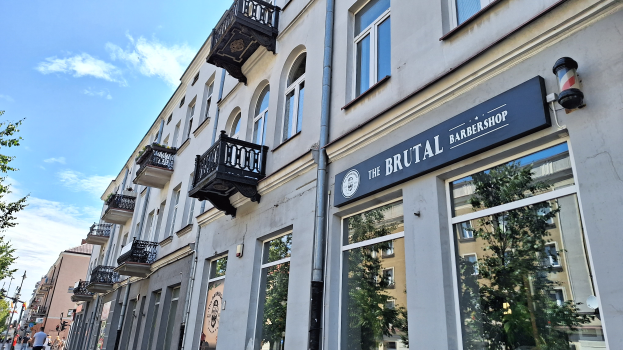 Eine Straßenszene in Berlin, Deutschland, mit einem Friseurladen, Gebäuden mit Fenstern, Geländern, Balkonen und Namensschildern, Bäumen, Laternenmasten und Menschen, die unter einem bewölkten Himmel auf der Straße gehen.