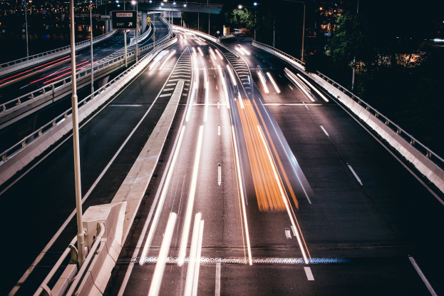 Langzeitbelichtung eines nächtlichen Highways mit Lichtspuren von fahrenden Fahrzeugen, beleuchteten Geländern, Pfosten, Planken, Straßenlaternen, Bäumen und Gebäuden im Hintergrund.