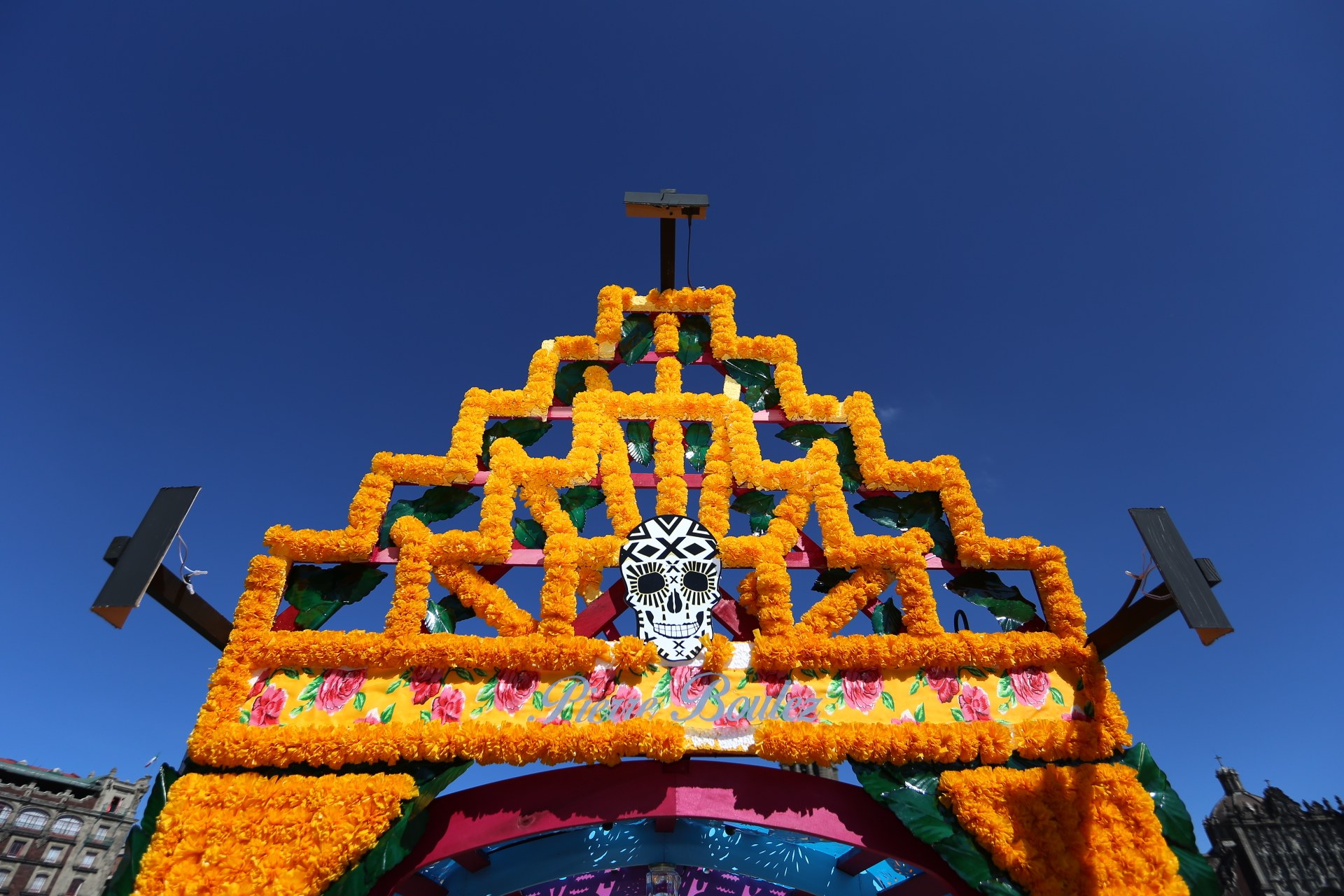 Ein Día de los Muertos-Bogen, geschmückt mit Blumen und einem Schädel, steht in Mexiko-Stadt vor Gebäuden unter einem klaren blauen Himmel.