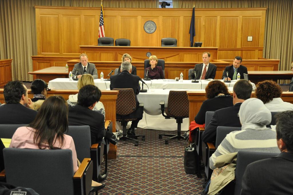 Ein Versammlungsraum mit einer Gruppe sitzender Menschen, zwei fremde Flaggen, ein oranger Schreibtisch mit Wasserflaschen und einem Mikrofon.
