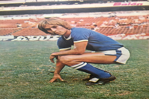 Ein Mann kniet auf einem Fußballfeld, umgeben von sitzenden Menschen und Stühlen mit Text darauf, trägt Schuhe.