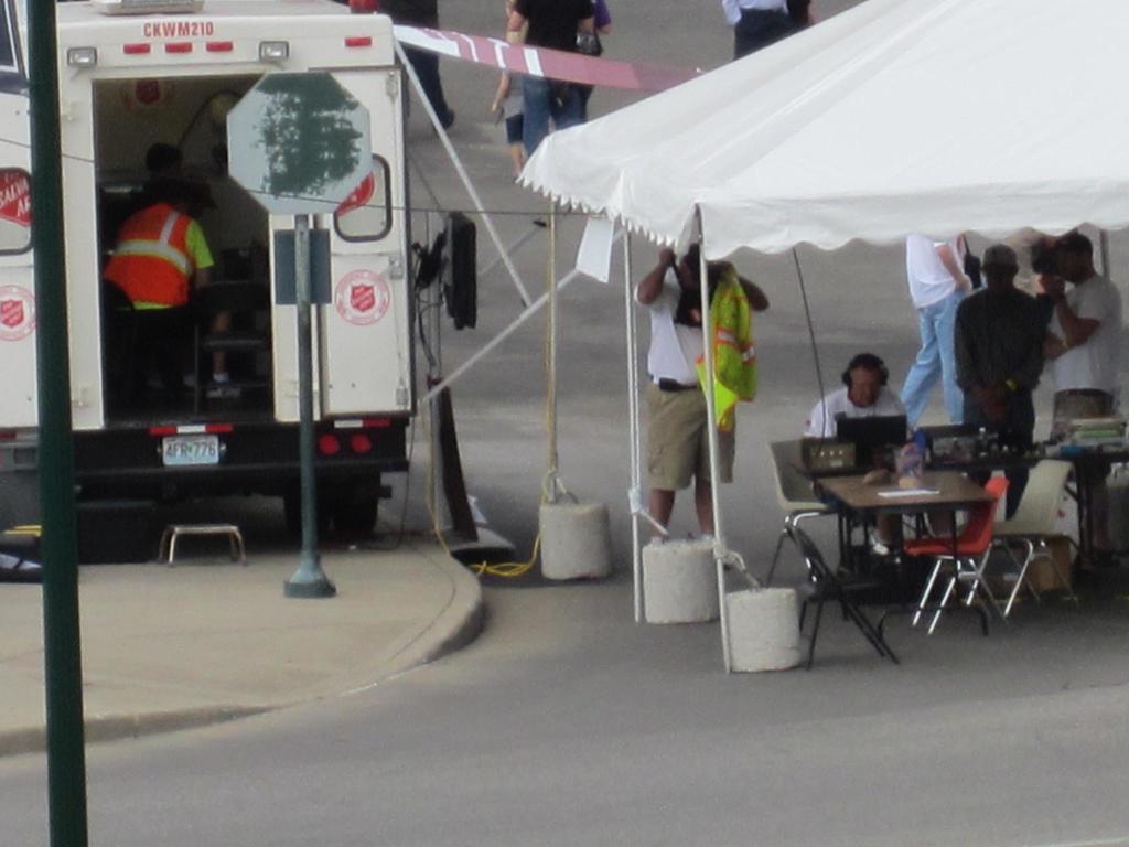 Ein Rettungswagen steht auf der linken Seite einer Stra√üe, w√§hrend auf der rechten Seite ein Zelt viele Menschen sch√ľtzt, mit einem Tisch und einem Stuhl in der N√§he.