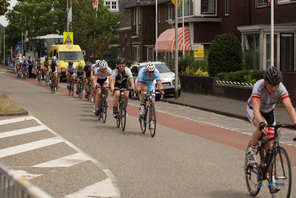 Menschen auf Fahrrädern mit Helmen, mit parkenden Autos auf der Straße und Fahnen an Masten, mit Bäumen und Gebäuden im Hintergrund.
