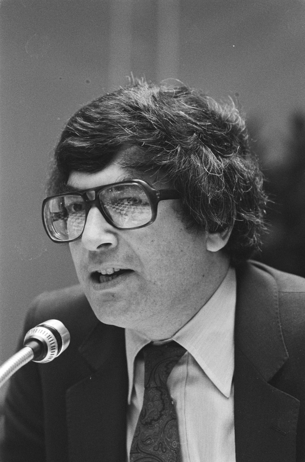 Ein Mann im Anzug und Krawatte, der eine Brille tr├Ągt, spricht ernst in ein Mikrofon, dargestellt in Schwarz-Wei├č.