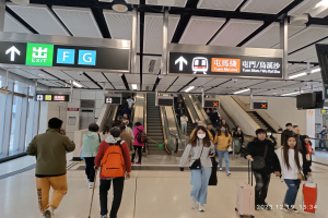 Menschen mit Gepäck durch einen geschlossenen Flughafen mit Rolltreppen, beleuchteten Schildern und Glaswänden im Hintergrund.