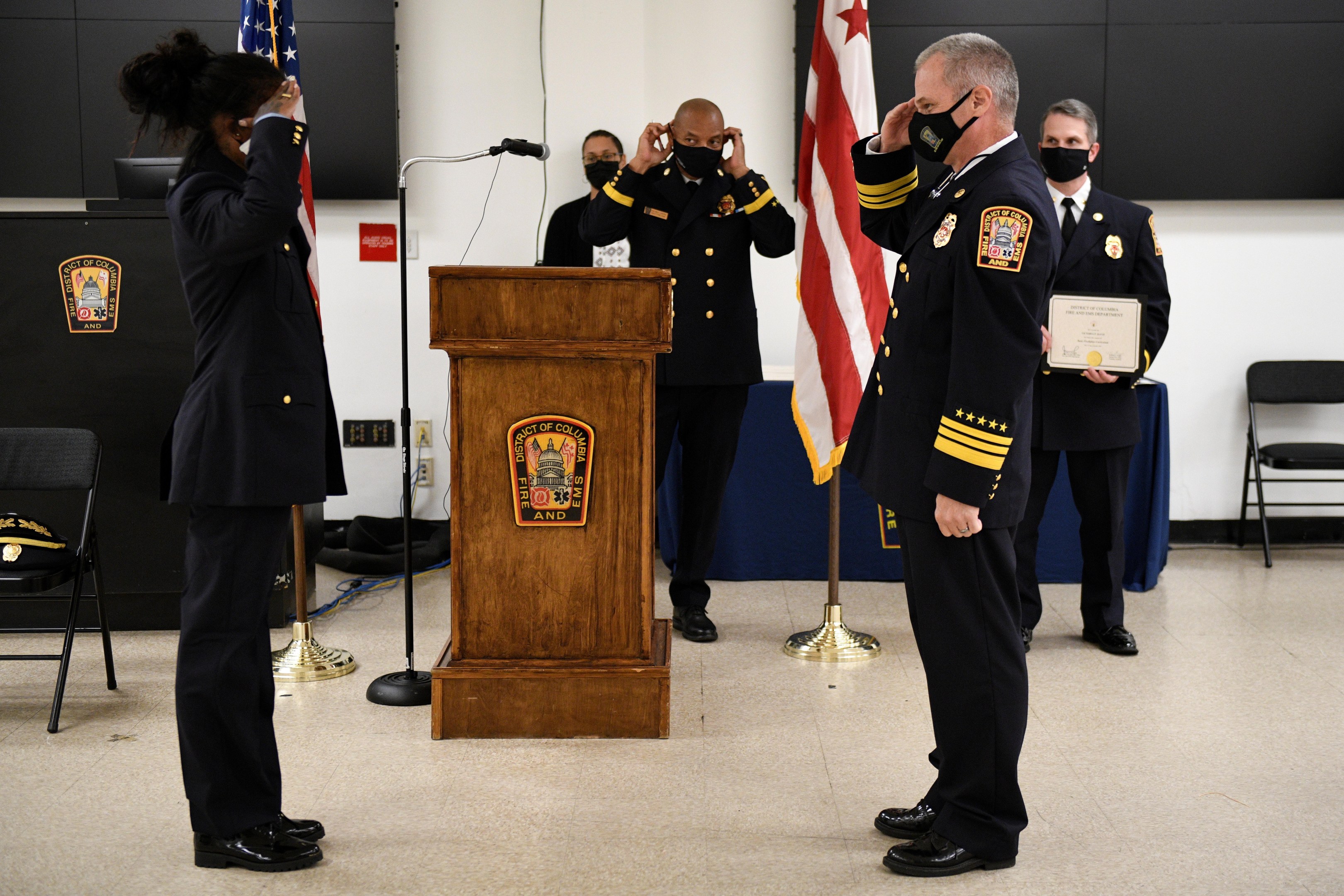 Gruppe uniformierter Personen auf einem Podium während einer Vereidigung eines neuen Feuerwehrchefs, der eine Urkunde hält, mit Flaggen und Stühlen im Hintergrund.