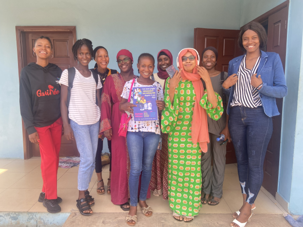 Gruppe von Frauen, die strahlen und Bücher in der Hand halten, vor einem Schulgebäude in Nigeria, mit einem drapierten Tuch auf dem Boden und Türen im Hintergrund.