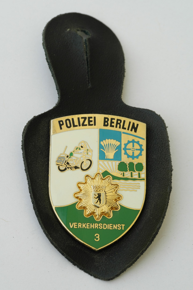 Ein schildförmiges Polizeibadge mit "Polizei Berlin" in schwarzer fetter Schrift auf einem blauen Hintergrund mit einem weißen Stern und Rahmen, das auf einer Oberfläche liegt.