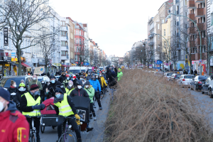 Eine große Gruppe von Menschen mit Masken und Sicherheitswesten fährt Fahrräder eine straße entlang, gesäumt von Bäumen, Gebäuden, Laternenmasten und Texttafeln, während Fahrzeuge die Straße teilen und trockenes Gras die rechte Seite saumt, unter einem klaren blauen Himmel.