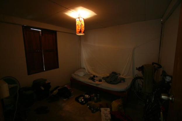 Ein Zimmer mit einem Bett in der Mitte, ein Fahrrad auf der rechten Seite, ein Fenster auf der linken Seite, ein Tischventilator, Taschen und Flaschen unten, mit Licht von oben.