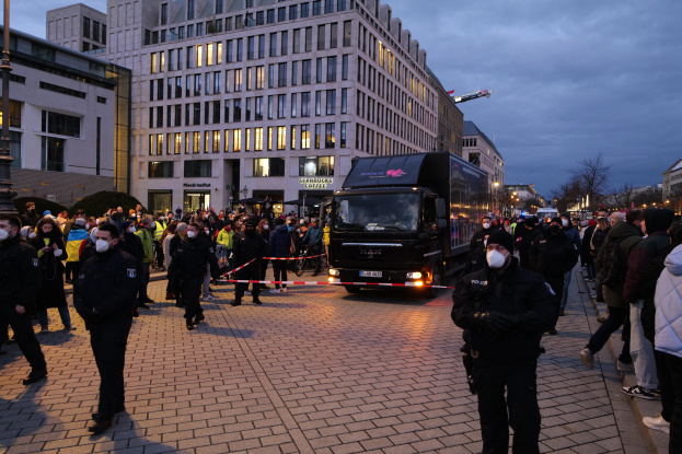 Eine Gruppe von Menschen steht vor einem Lkw auf einer Straße, umgeben von Gebäuden, Laternen, Bäumen und einem bewölkten Himmel, wobei einige Mützen und Masken tragen und ein Band mit einem Stab im Vordergrund zu sehen ist.
