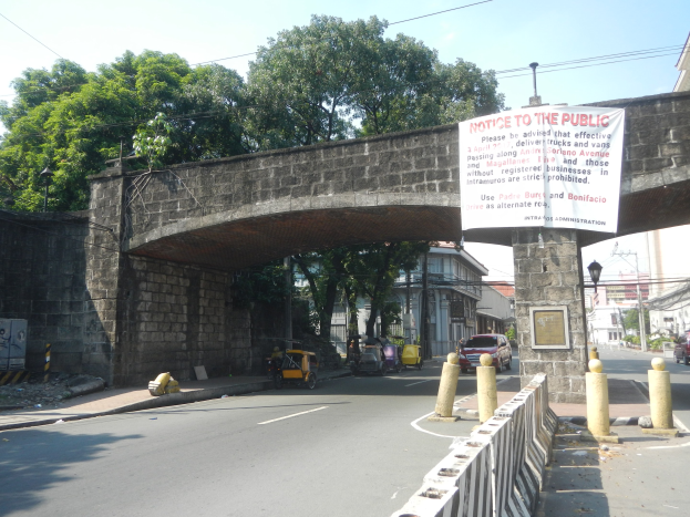 Öffentlicher Hinweisschild auf einer Brücke über einer Straße mit Fahrzeugen, einem Geländer auf der rechten Seite, Gebäuden, Bäumen, Strommasten, Drähten und Himmel im Hintergrund.