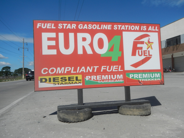 Eine Tankstelle mit einem "Euro4 Fuel"-Schild im Vordergrund, Fahrzeuge auf der Straße, Strommasten mit Drähten, Bäume, ein Gebäude auf der rechten Seite und ein sichtbarer Himmel.