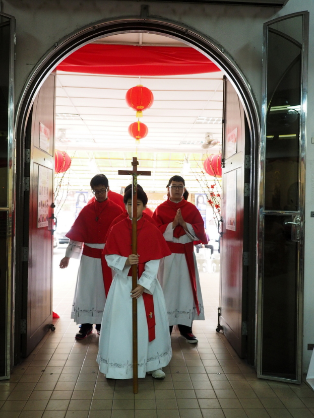 Menschen, die mit einem Kreuz in einer Kathedrale gehen, mit Türen links und rechts und Blumen und dekorativen Elementen im Hintergrund.